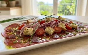 bresaola-marinata-mediterranea-finocchi-pomodorini-crostini Bresaola stesa su un piatto piano marinata con limone e arancia, coperta da un'insalata croccante di finocchi rossi affettati finissimi, pomodorini ciliegino a metà e crostini di pane tostato al timo.