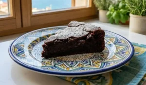 Una fetta di Torta Caprese ricca e umida al cioccolato e mandorle con zucchero a velo, servita su un piatto di ceramica vietrese colorata in una luminosa cucina mediterranea