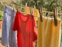 Primo piano di una trappola per moscerini della frutta fai-da-te: un barattolo di vetro con aceto di mele all'interno, coperto da pellicola trasparente con piccoli fori e fissato con spago, su un tavolo di legno. Sullo sfondo un cesto di pesche e banane.
