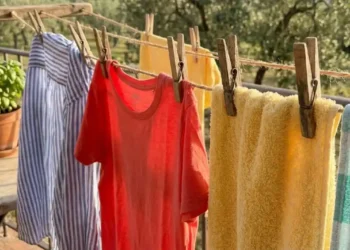 Primo piano di una trappola per moscerini della frutta fai-da-te: un barattolo di vetro con aceto di mele all'interno, coperto da pellicola trasparente con piccoli fori e fissato con spago, su un tavolo di legno. Sullo sfondo un cesto di pesche e banane.