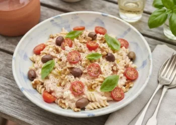 Piatto di pasta fredda cremosa con pomodorini, ricotta, noci e basilico pronto da servire su un tavolo in legno.