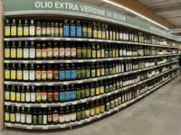 Ampio scaffale di un supermercato ben fornito con numerose marche e bottiglie di olio extravergine d'oliva in vetro scuro.